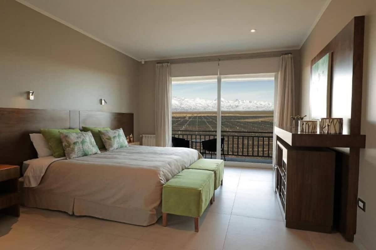 A bedroom with a large bed, two green ottomans, a wooden nightstand, a dresser, and a balcony with a view of snow-capped mountains.