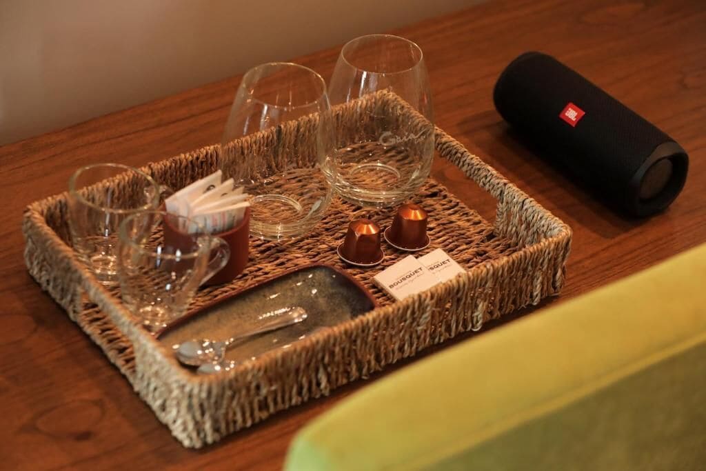 A tray with glasses, a bottle, and a small dish on a woven mat, accompanied by a black cylindrical object.