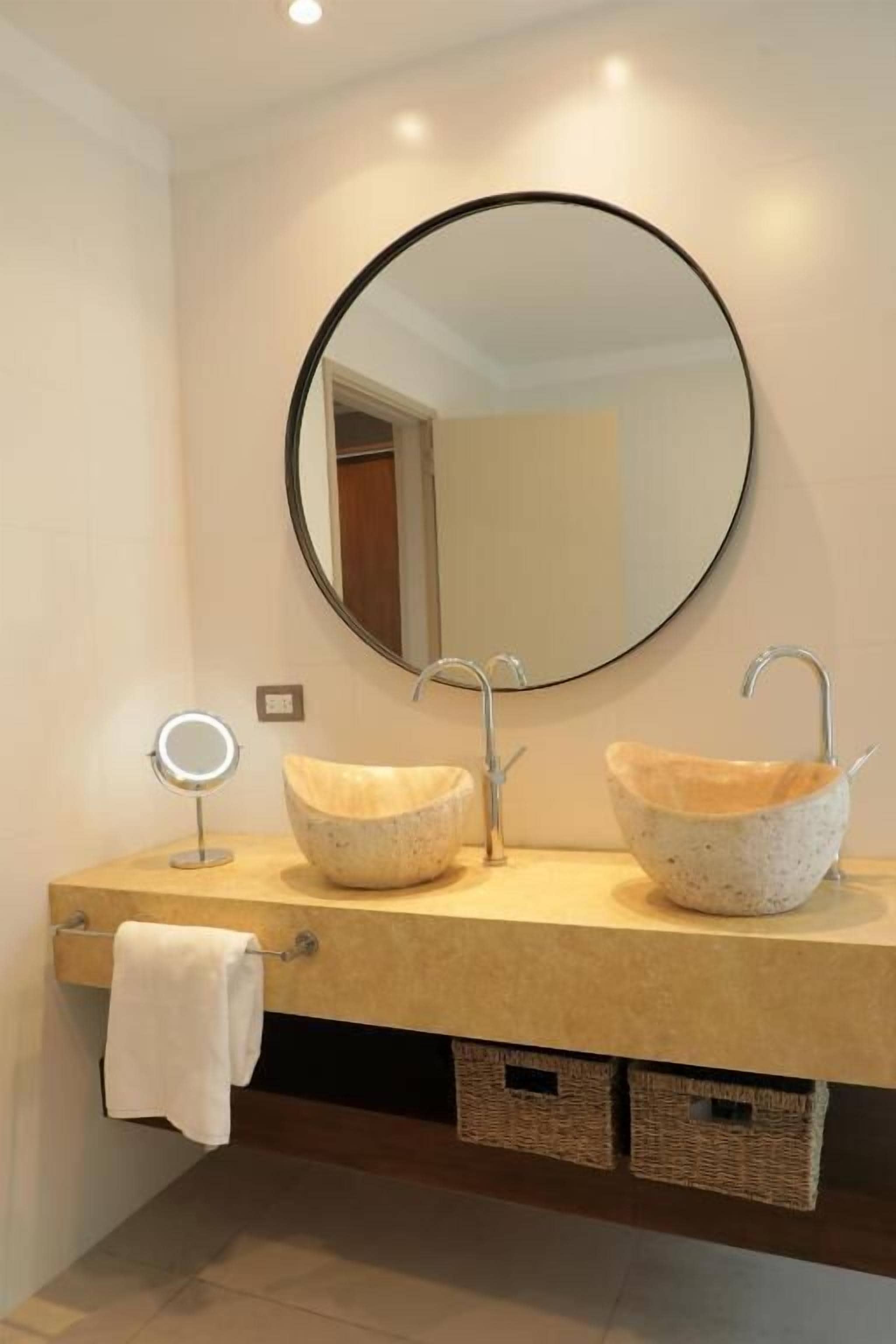 Un baño con un espejo redondo, dos lavamanos con textura similar a la piedra y un perchero para toallas.
