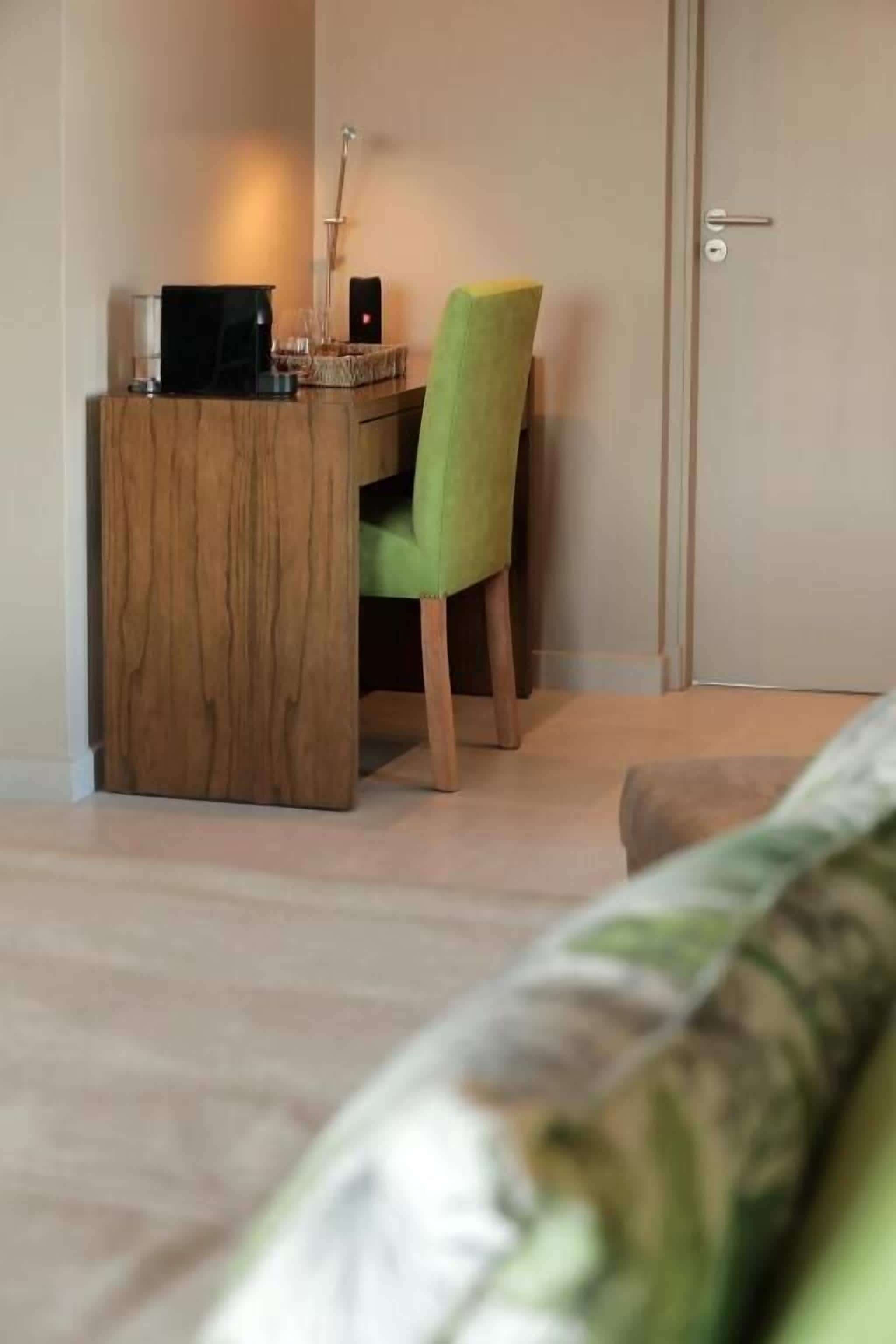 A room with a wooden desk, a green chair, a bed with a patterned bedspread, and a door.