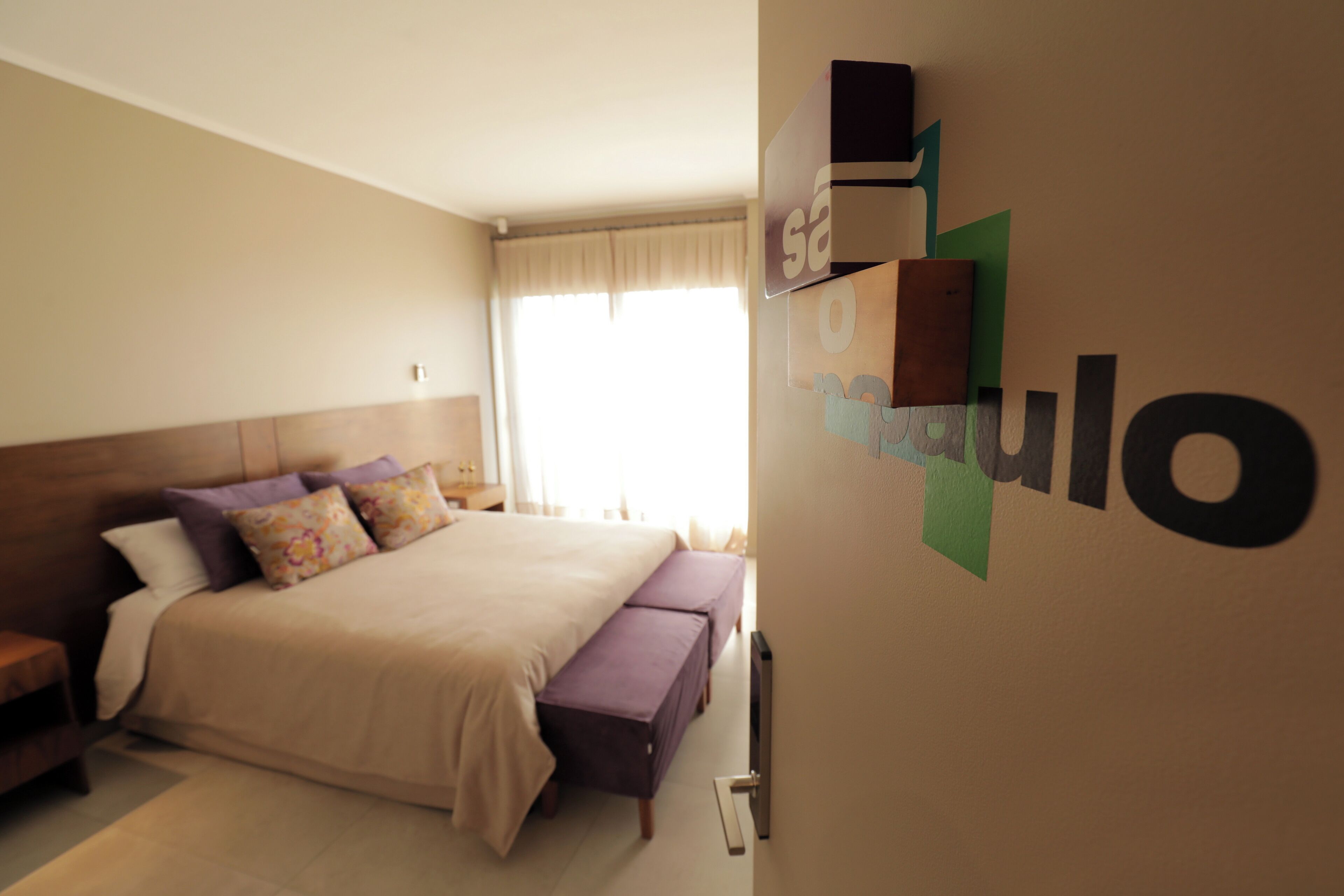 Quarto com cama, um banquinho, mesa de cabeceira e prateleira na parede.