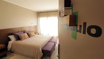 Quarto com cama, um banquinho, mesa de cabeceira e prateleira na parede.