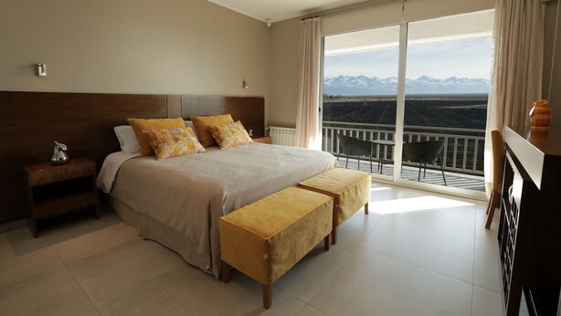A bedroom with a large bed, a nightstand, a bench, and a balcony with a view of mountains. - Gaia Lodge (Tupungato)