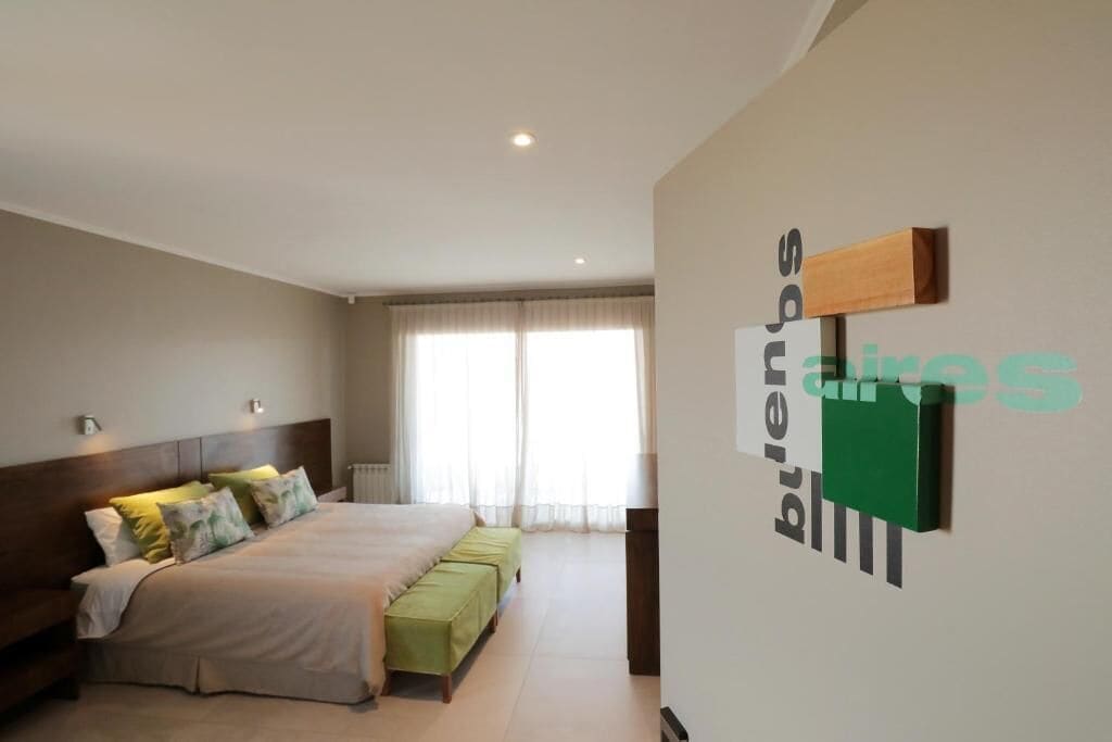 Une chambre à coucher comprenant un lit, des tables de chevet, une fenêtre avec des rideaux et une œuvre d’art accrochée au mur.