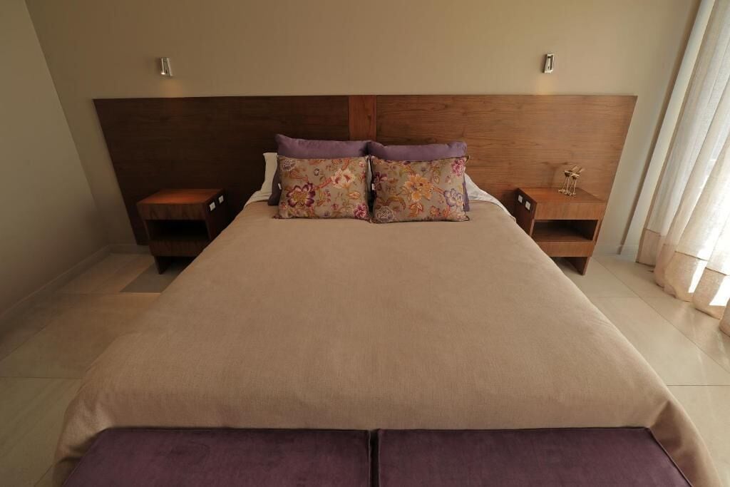 Una cama bien tendida con un juego de almohadas florales, cabecera de madera y mesitas de noche con lámparas.