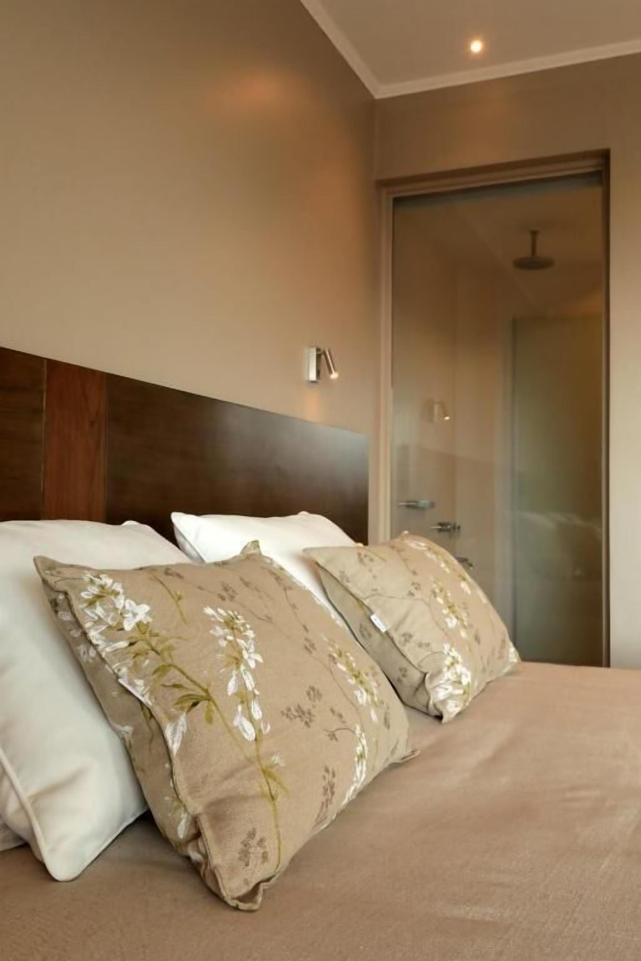 A hotel room with a bed, two pillows, a nightstand, a wall-mounted lamp, and a sliding glass door leading to a shower area.