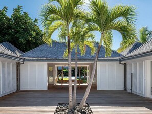 A covered outdoor area with a wooden deck, two palm trees, and a view of a house with a dark roof and white garage doors.