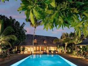 A poolside area with a wooden deck, lounge chairs, and a building with a thatched roof.
