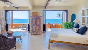 A bedroom with a bed, a wooden nightstand, a wicker chair, a table, and a view of the ocean.