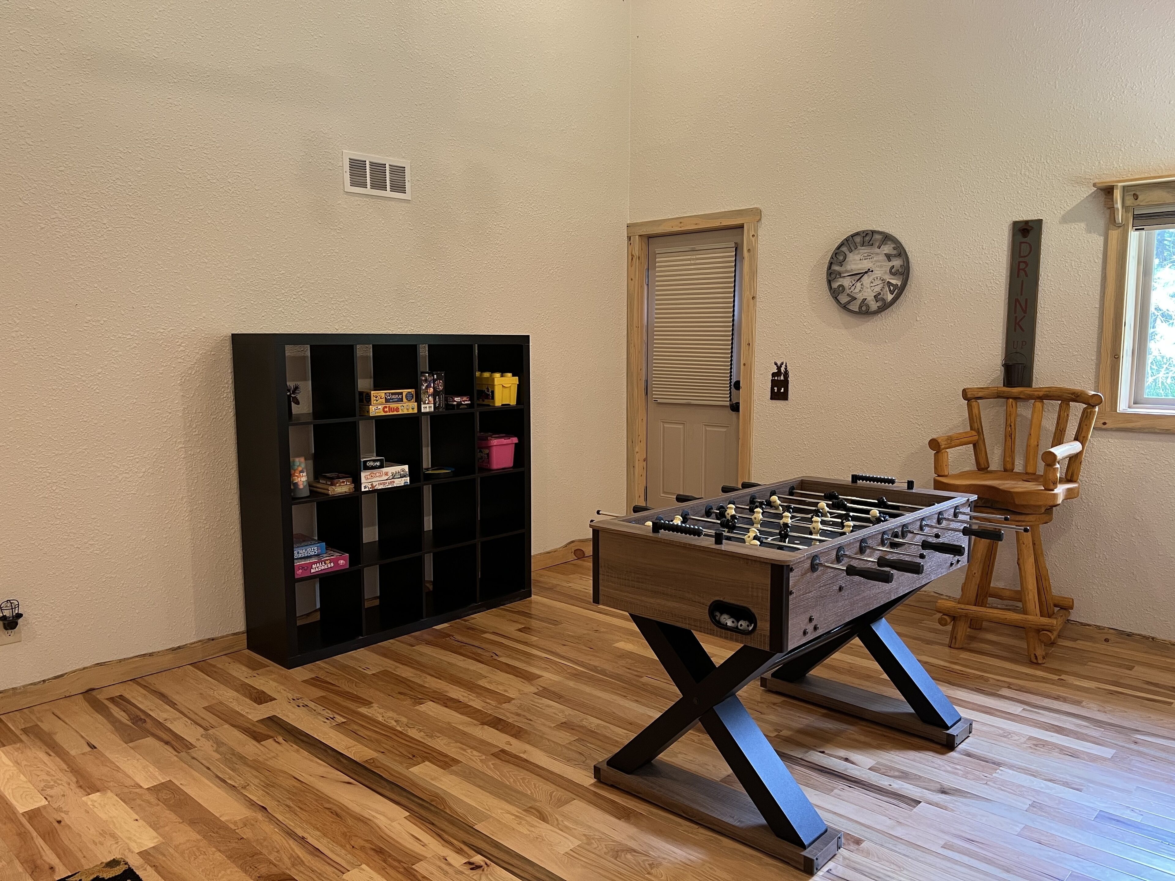 Ein Zimmer mit Holzboden, einem Kicker, einem schwarzen Regal mit diversen Gegenständen, einem Holzstuhl, einer Tür und einer Wanduhr.
