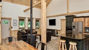 Un interior rústico con techo de madera, chimenea, zona de comedor con mesa y sillas de madera, cocina con una isla grande y un televisor montado en la pared.