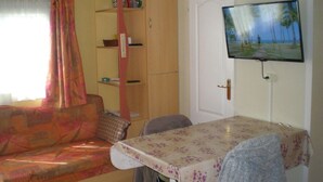 Un espace de vie compact comprenant un canapé, une table à manger recouverte d’une nappe à motifs floraux, un téléviseur fixé au mur et une armoire.