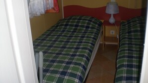 Une petite chambre avec deux lits simples, chacun recouvert d’une couverture à carreaux. On y trouve une table de chevet avec une lampe, ainsi qu’une petite table dotée d’une prise électrique.