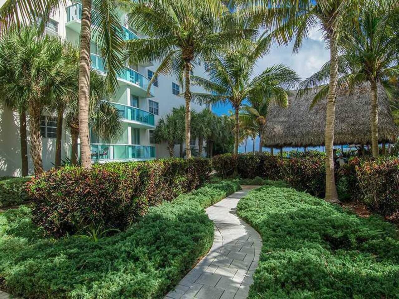 A pathway flanked by lush greenery and palm trees, leading to a modern building with balconies.