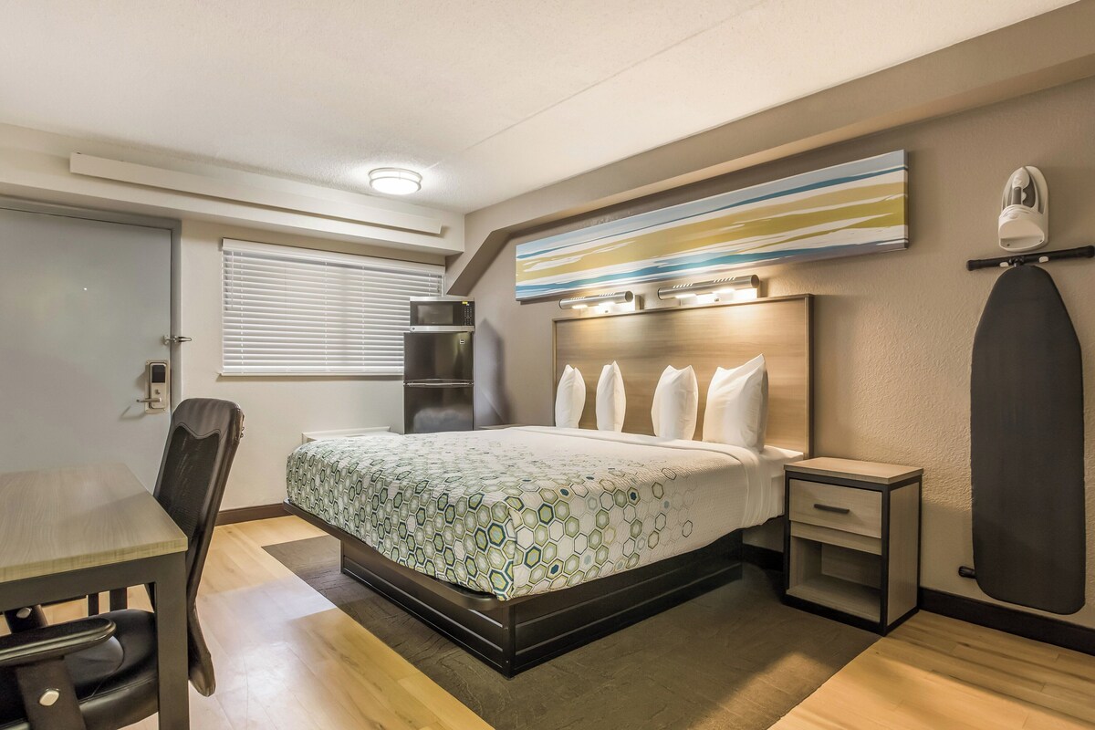 A modern hotel room with a large bed, a desk, a chair, a small bedside table, and a wall-mounted ironing board.