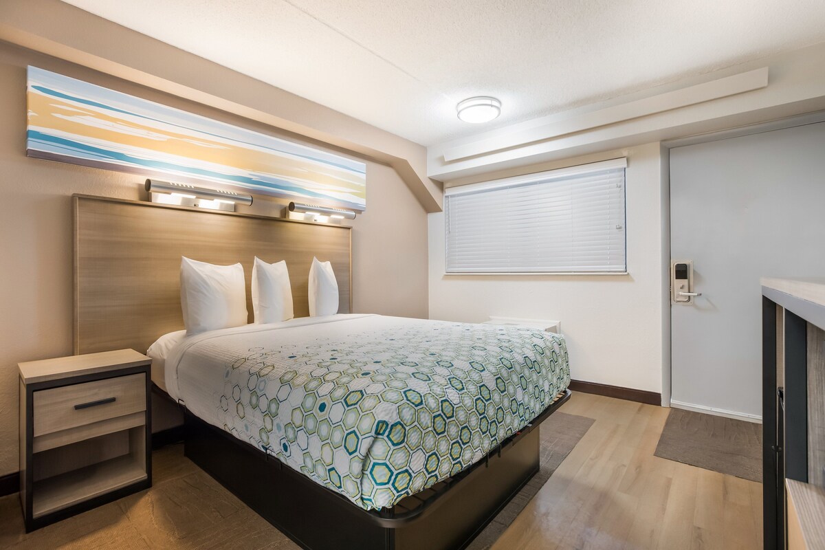 Una habitación de hotel moderna con una cama, mesitas de noche, cabecera, ventana con persianas y una lámpara en el techo.