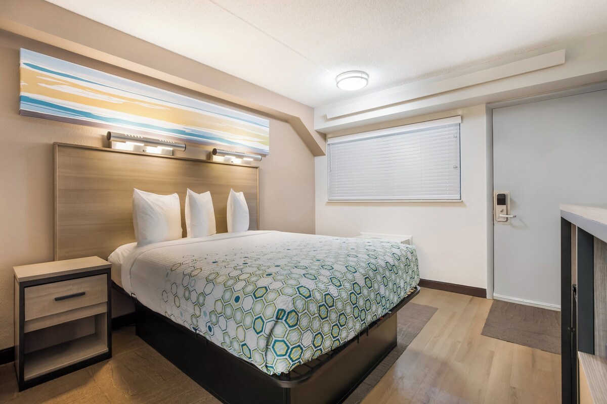 A modern hotel room with a bed, bedside tables, a headboard, a window with blinds, and a ceiling light.