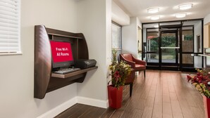 A modern hotel room with a built-in desk, a red sign indicating free Wi-Fi, a red chair, and a potted plant. - HomeTowne Studios By Red Roof Princeton – Ewing/ Lawrenceville (Lawrenceville)
