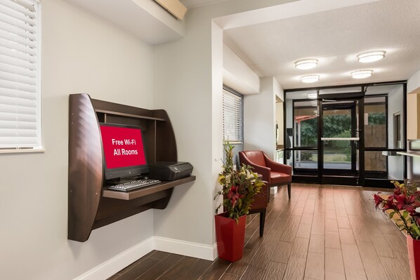 A modern hotel room with a built-in desk, a red sign indicating free Wi-Fi, a red chair, and a potted plant. - HomeTowne Studios By Red Roof Princeton – Ewing/ Lawrenceville (Lawrenceville)