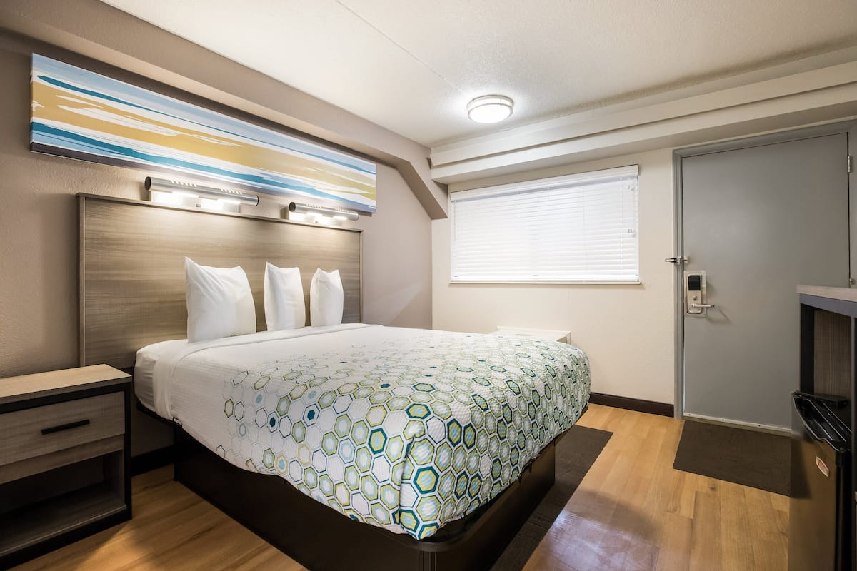 A modern hotel room with a large bed, a bedside table, a nightstand, a window with blinds, and a door.