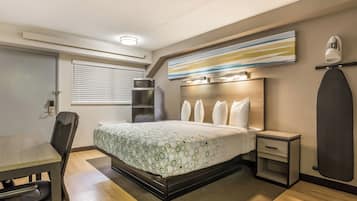 Habitación de hotel con cama, escritorio, silla, microondas, refrigerador, mesita de noche y tabla de planchar en la pared.