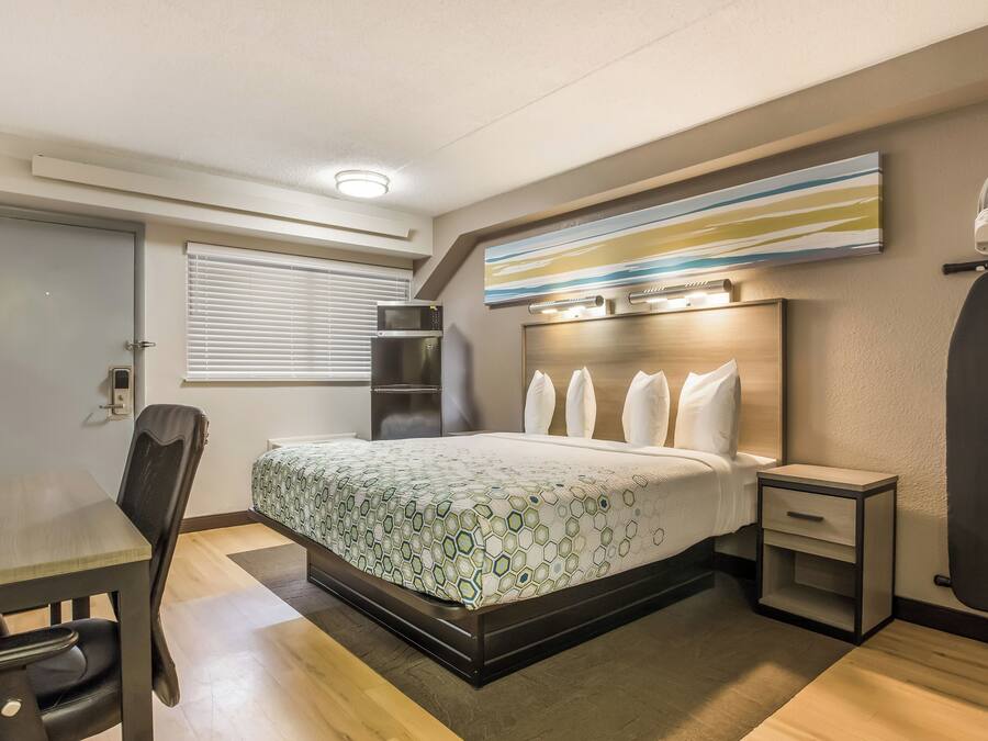 Ett hotellrum med en säng, ett skrivbord, en stol, en mikrovågsugn, ett kylskåp, ett nattduksbord och en väggmonterad strykbräda.