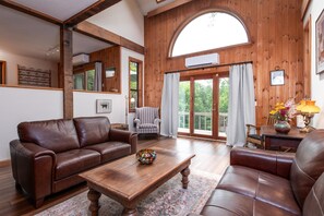 A spacious living room with a brown leather sofa, a wooden coffee table, and a striped armchair. A large window with a view of greenery is visible. - Romantic Retreat - Minutes from Stratton (Londonderry)