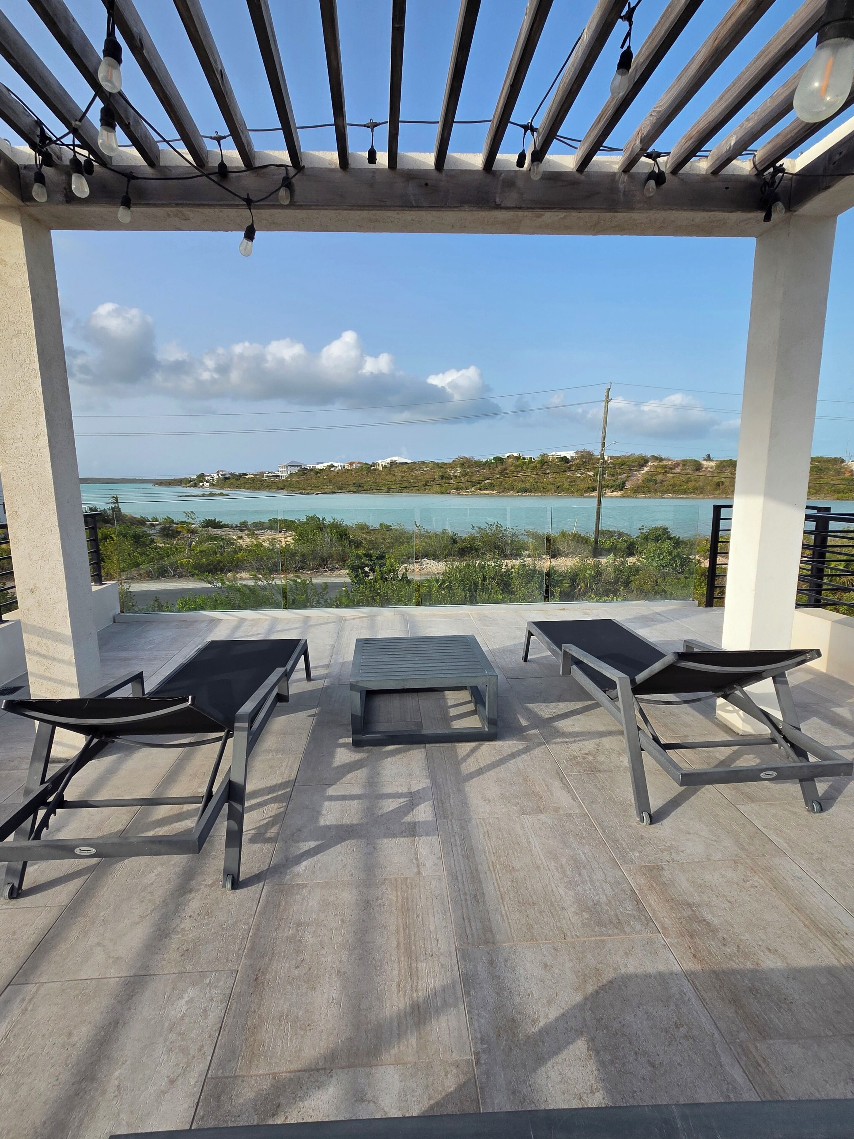 A covered outdoor seating area with two lounge chairs and a table, overlooking a coastal landscape.