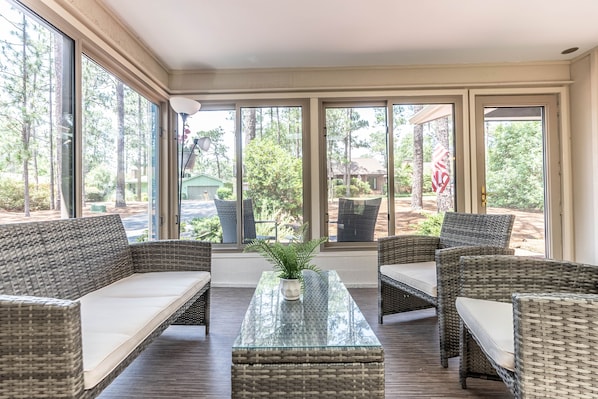 Relax in this inviting sunroom with a glass coffee table and wicker furniture, surrounded by views of a landscaped garden.