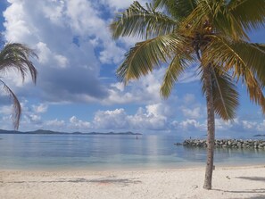 Una playa con agua cristalina, palmeras y montañas a lo lejos.