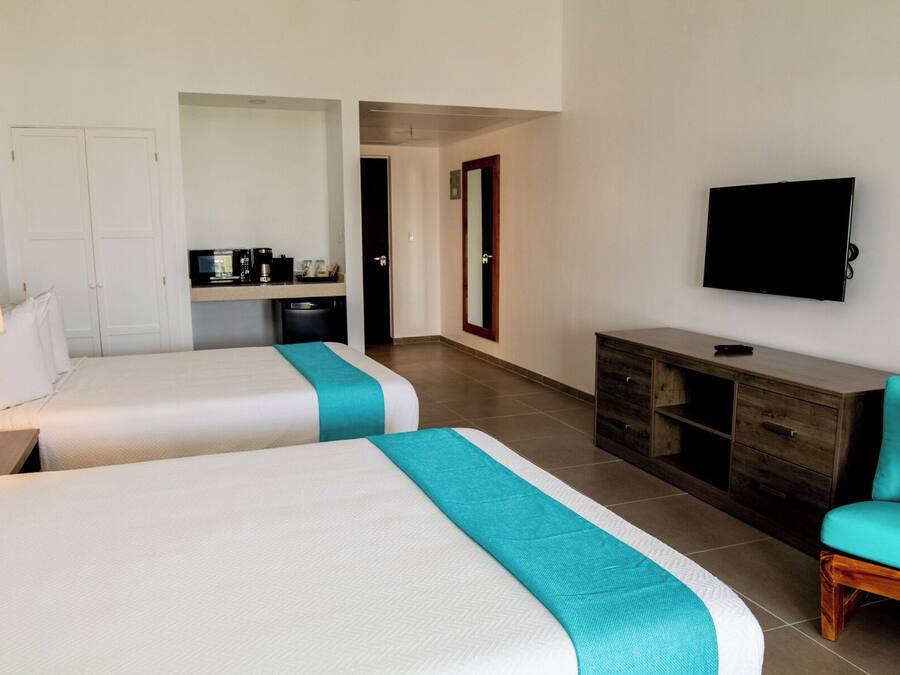 Quarto de hotel com duas camas, televisão, escrivaninha com computador e cozinha compacta.