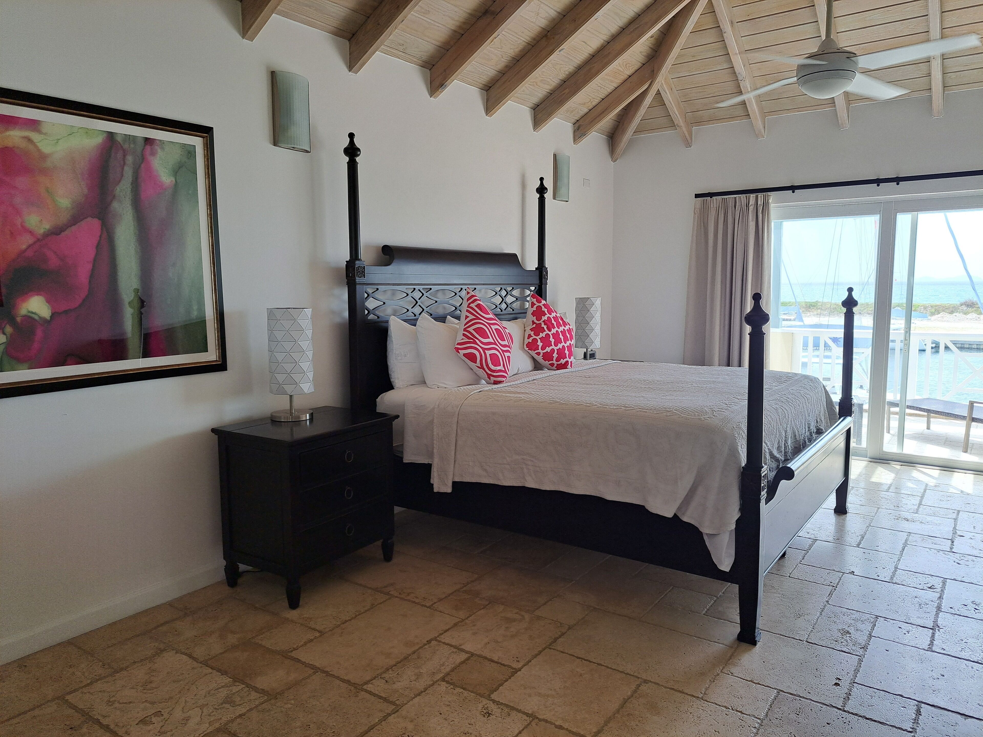 A bedroom with a bed, a nightstand, a painting, and a view of the sea through a large window.