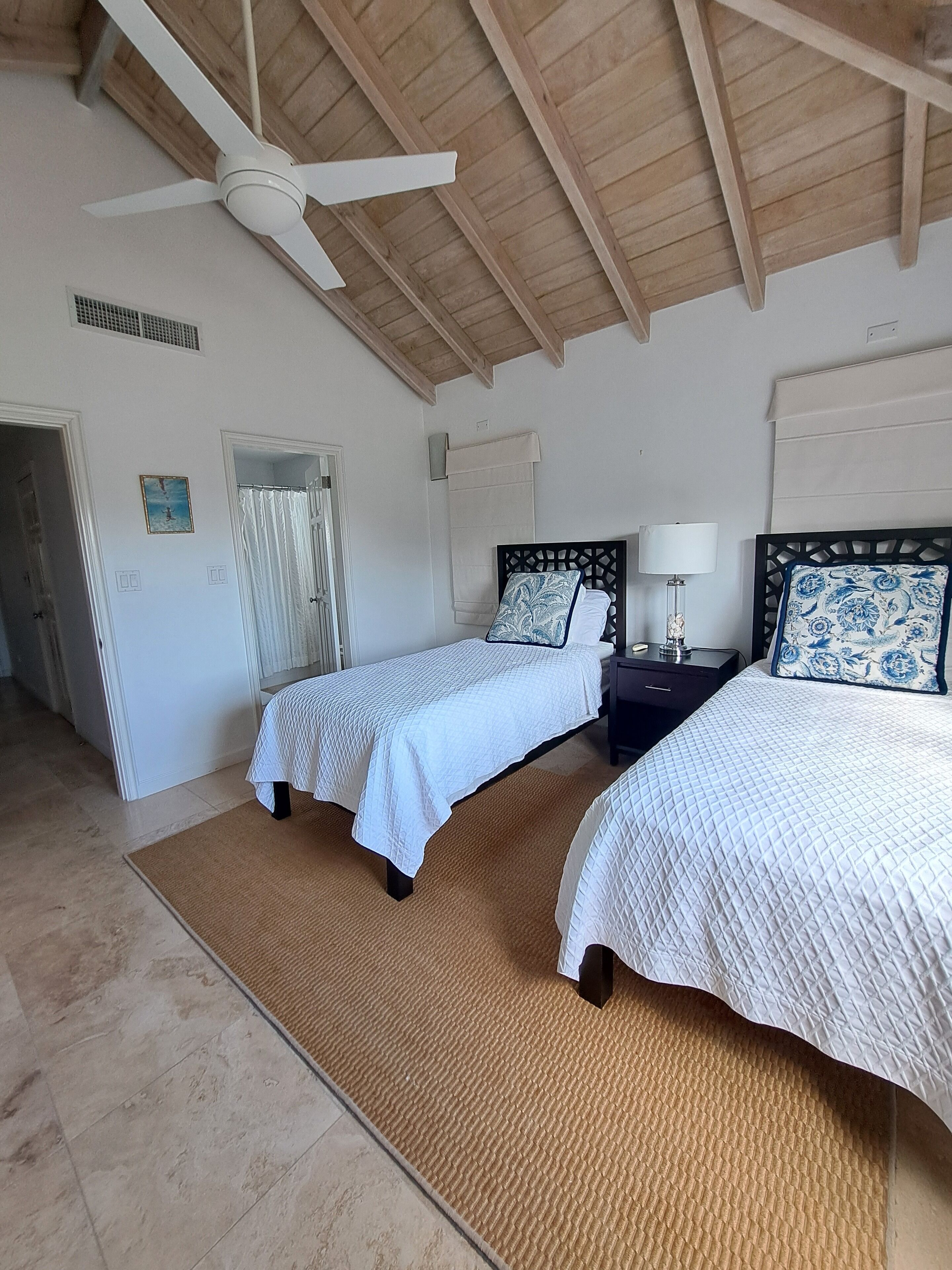 A bedroom with two beds, a ceiling fan, a wooden ceiling, and a view of the beach.