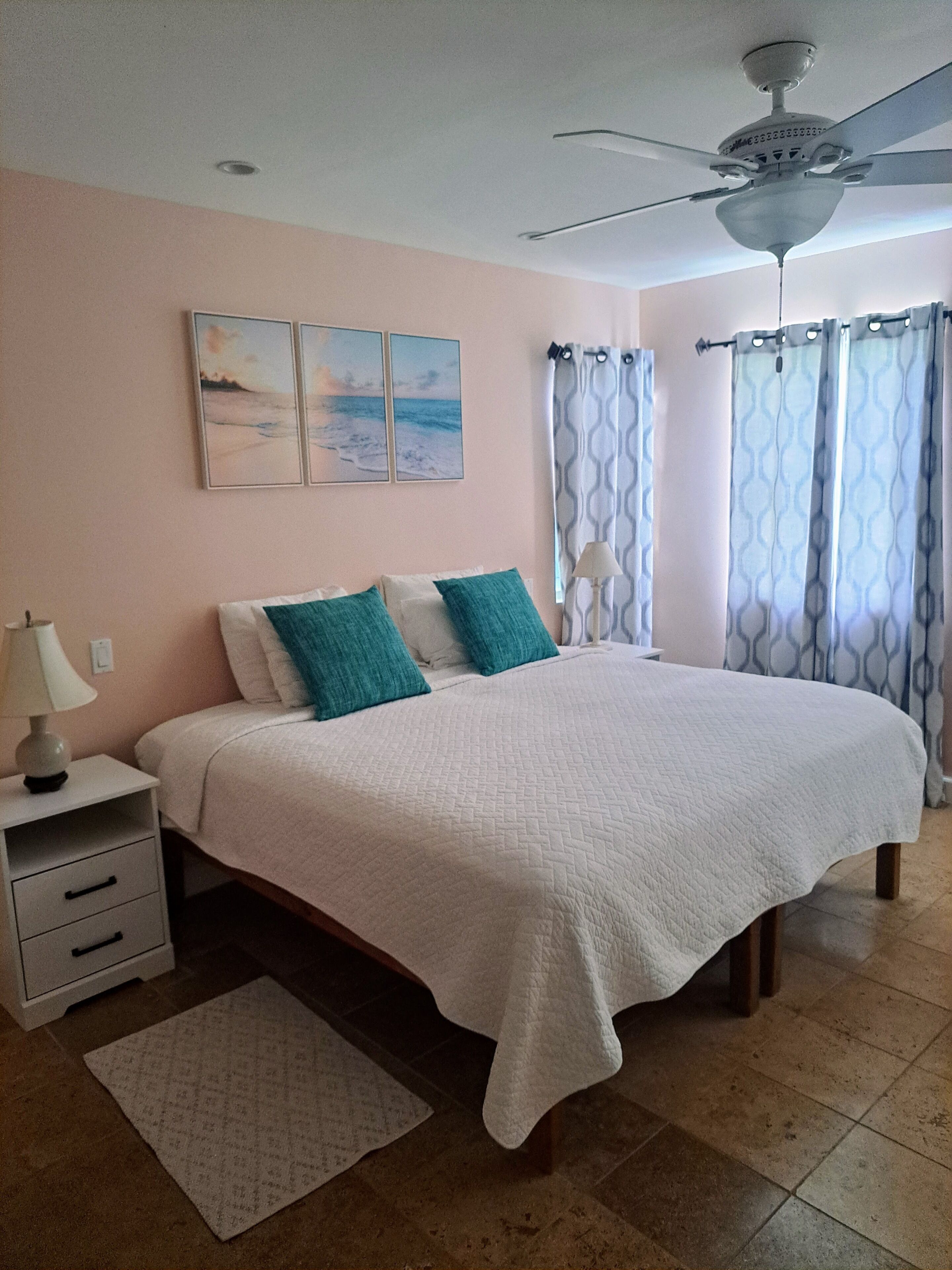 A bedroom with a bed, a nightstand, a lamp, a ceiling fan, and a window with curtains.