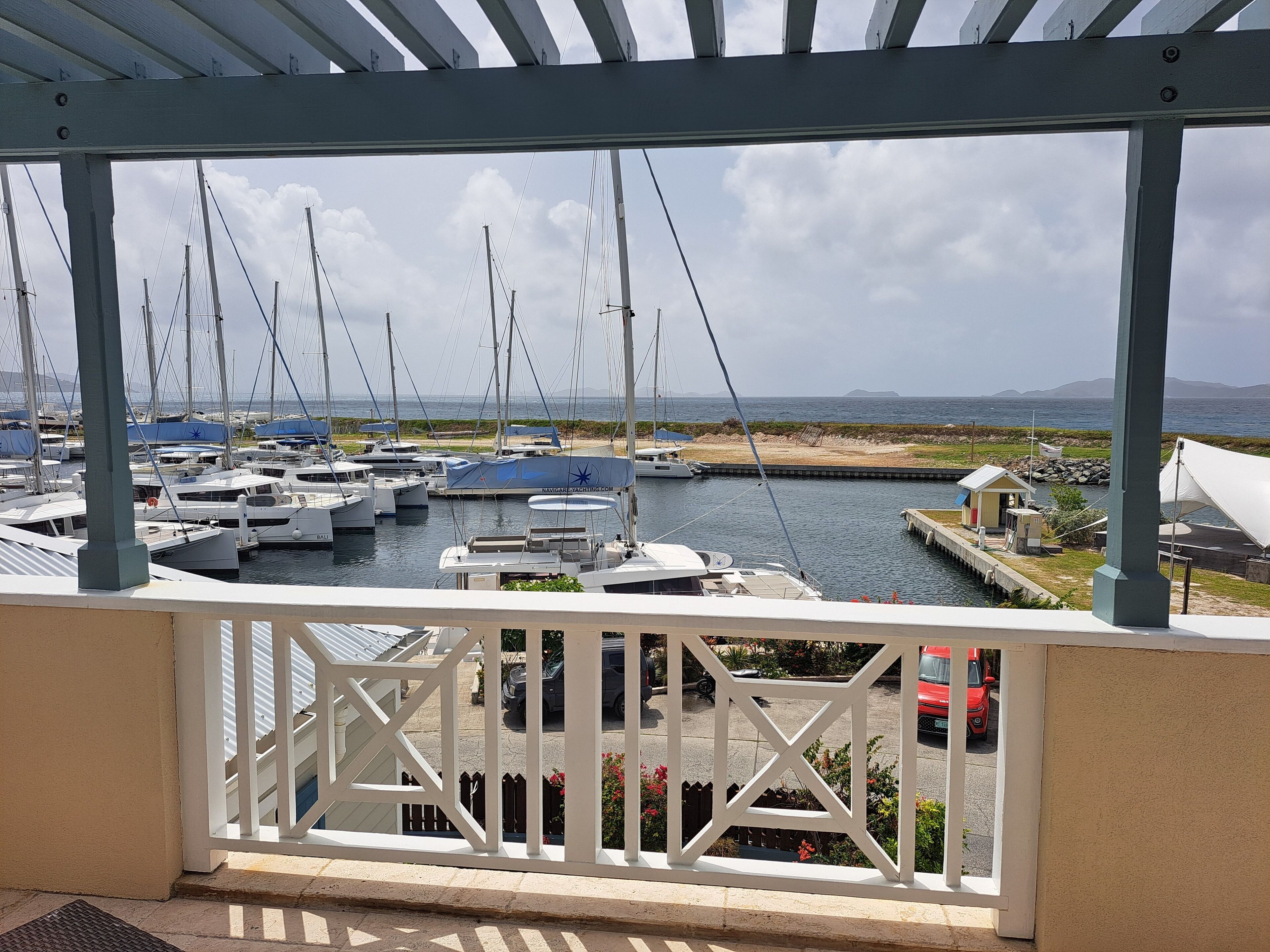 A marina with multiple boats docked, a covered area with a railing, and a view of the sea and distant land.