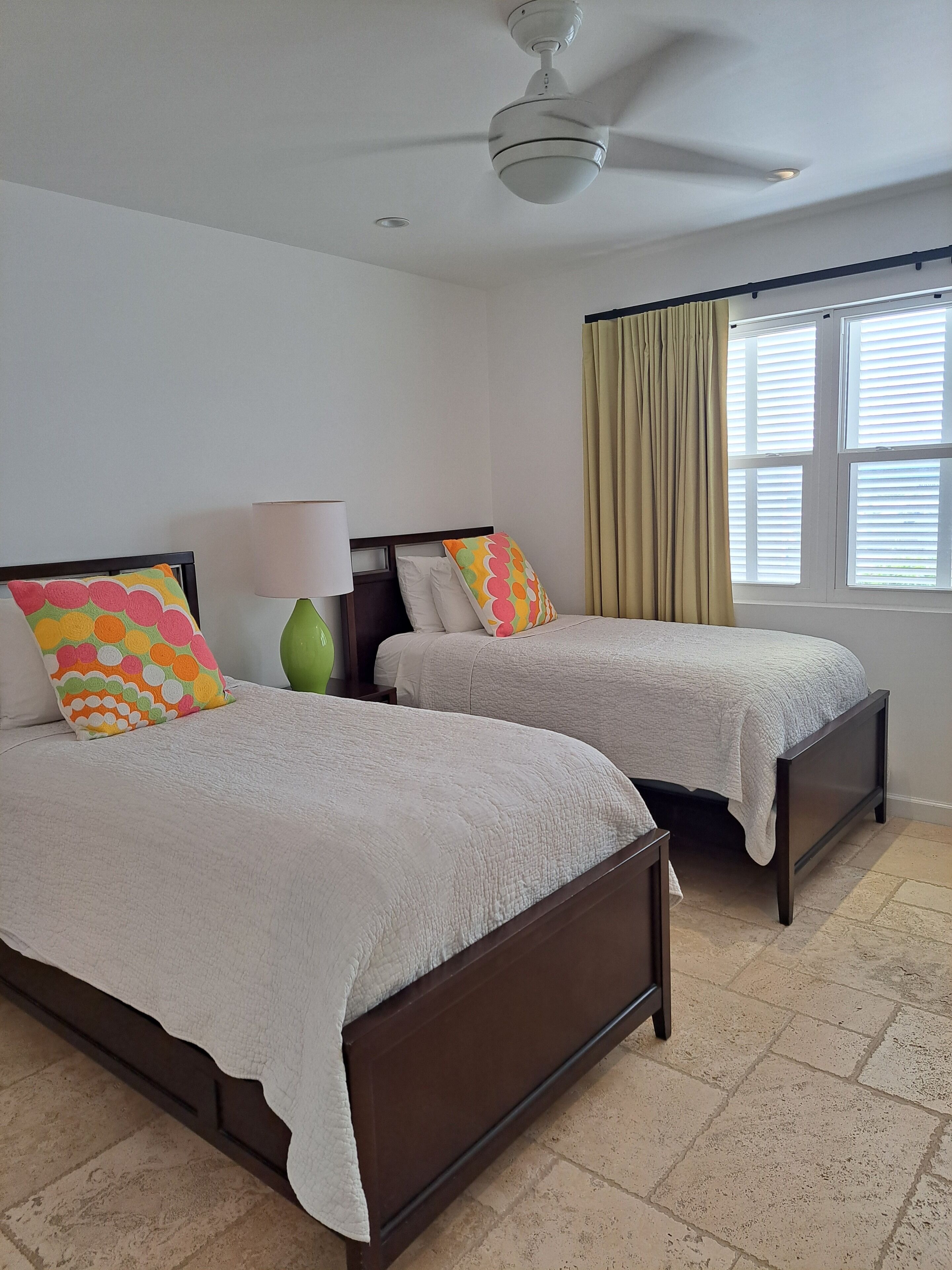 A hotel room with two beds, a ceiling fan, a window with curtains, and a lamp.