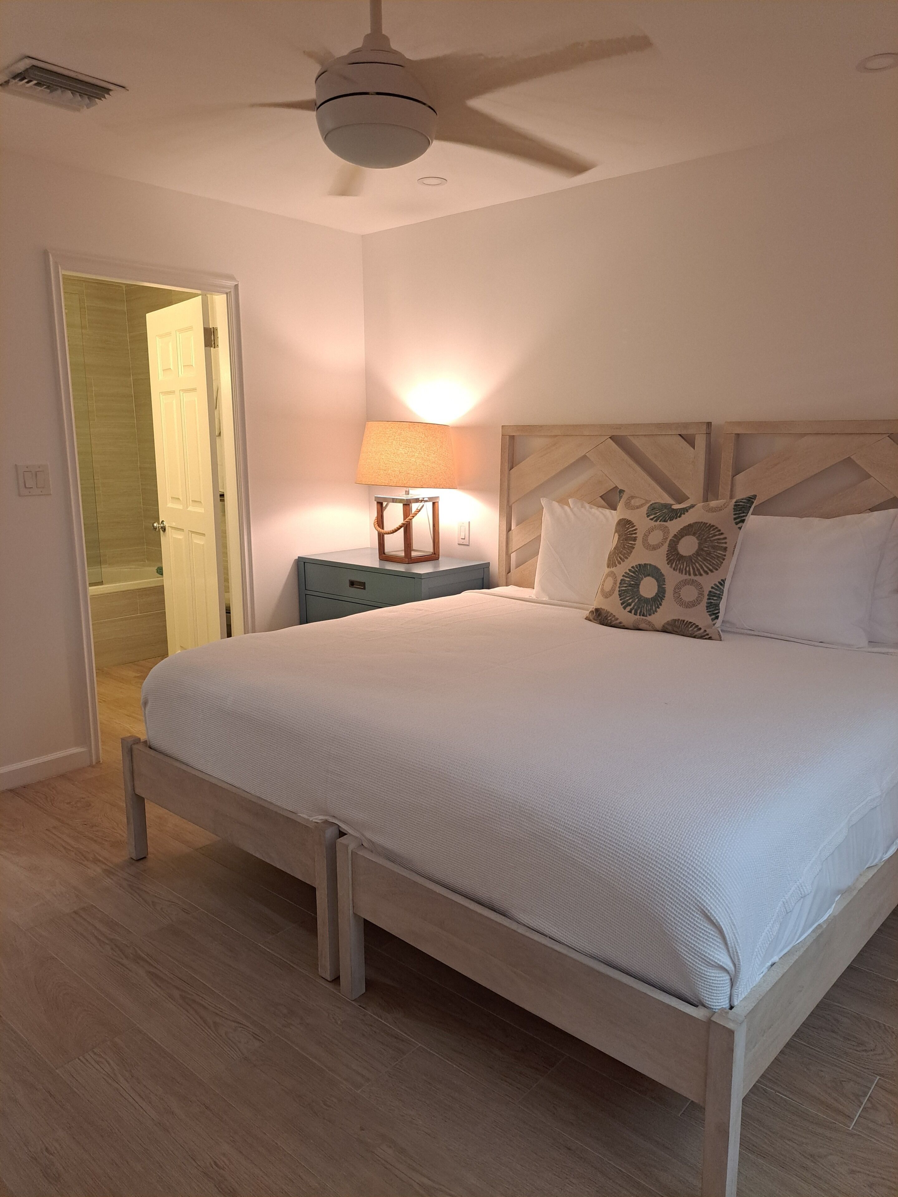 A bedroom with a bed, a nightstand, a lamp, a ceiling fan, and a door leading to a bathroom.
