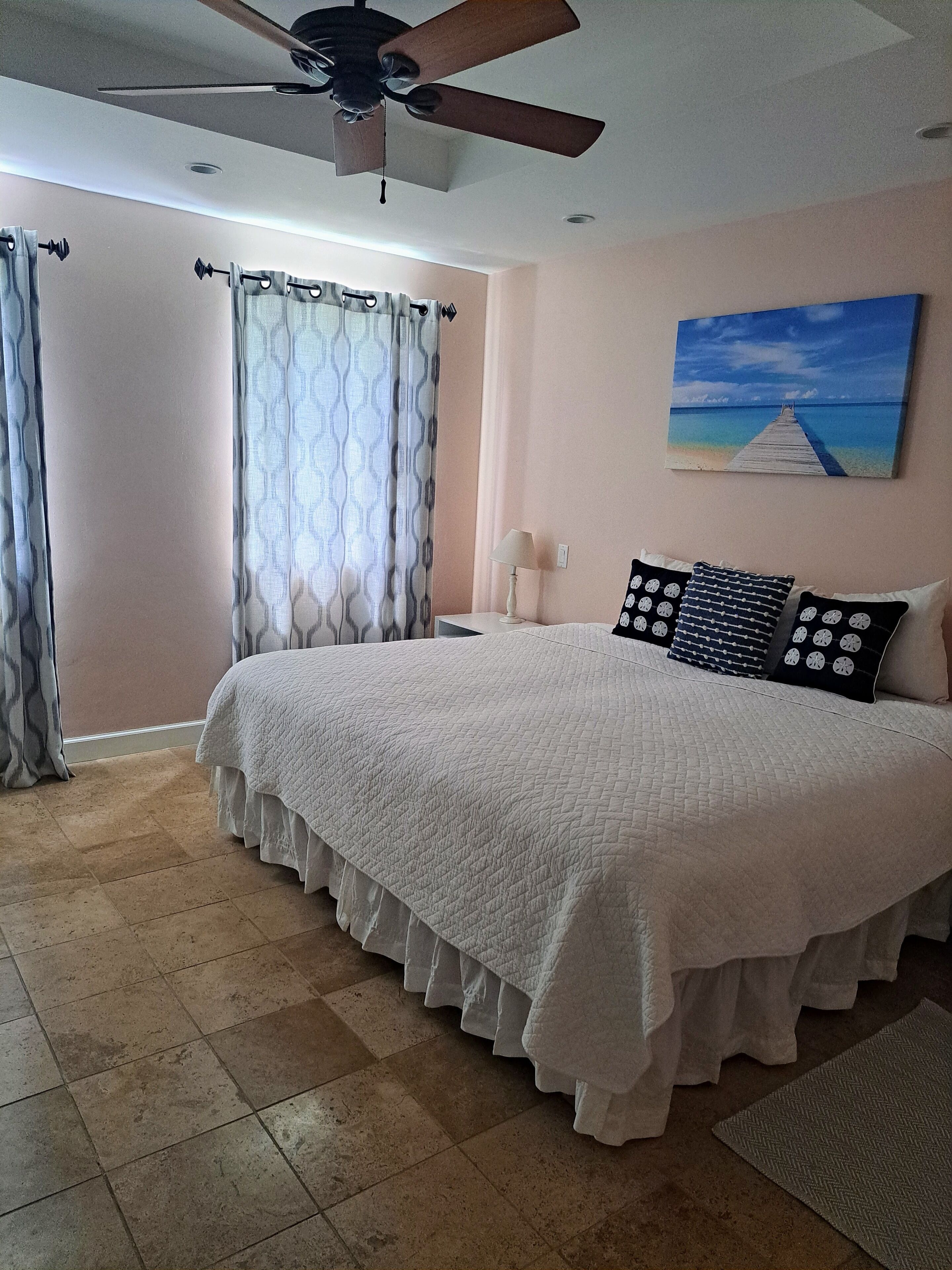 Un dormitorio con cama, cortinas, ventilador de techo, un cuadro y suelo de baldosas.