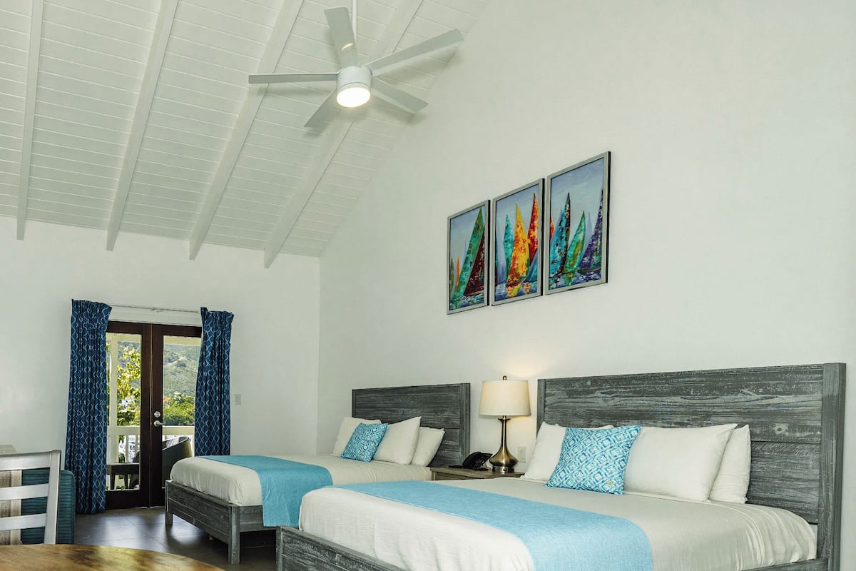 Une chambre d’hôtel avec deux lits, un ventilateur de plafond, une table de chevet avec une lampe et une porte donnant sur un espace extérieur.