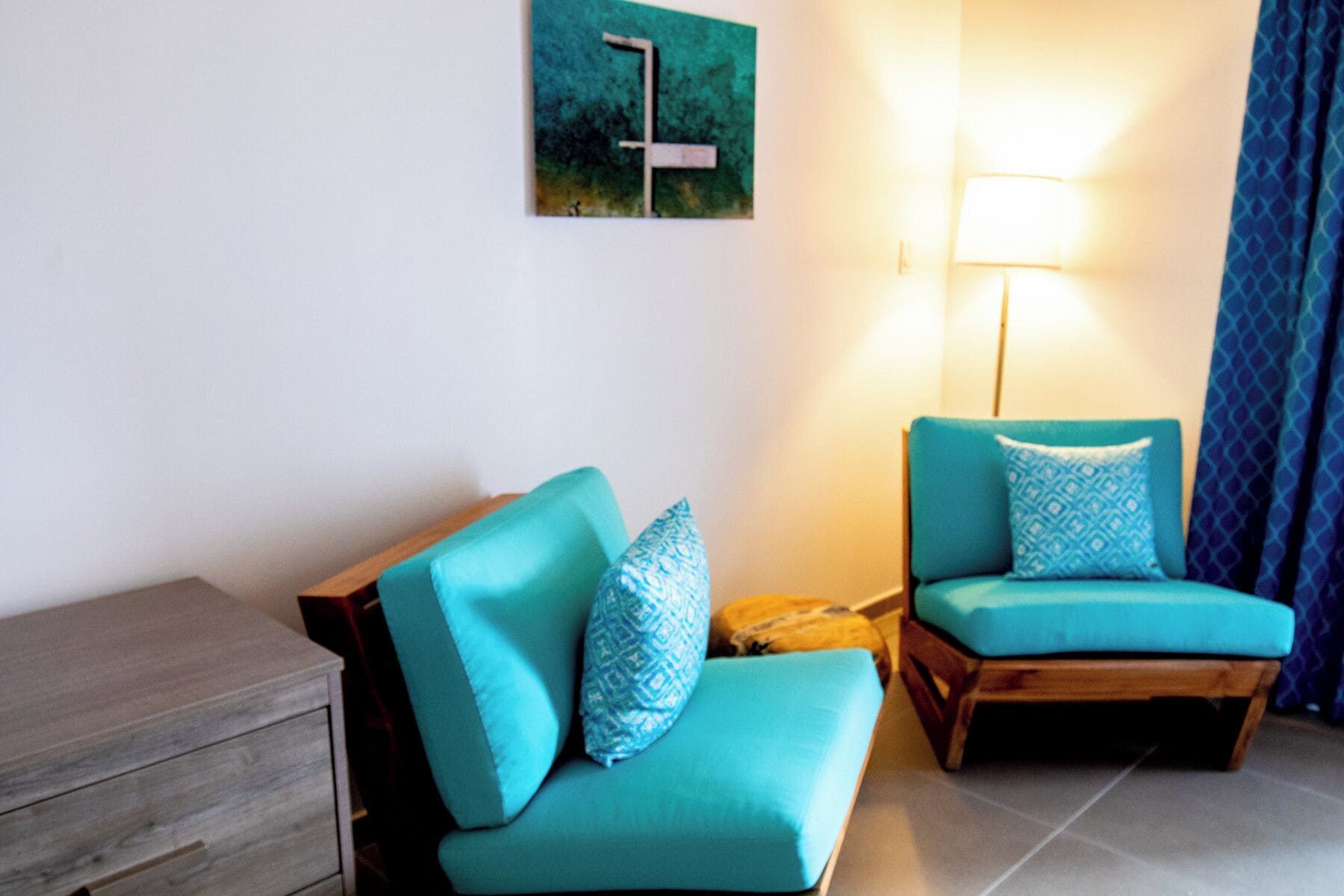 A room with two teal armchairs, a wooden side table, a floor lamp, and a painting on the wall.