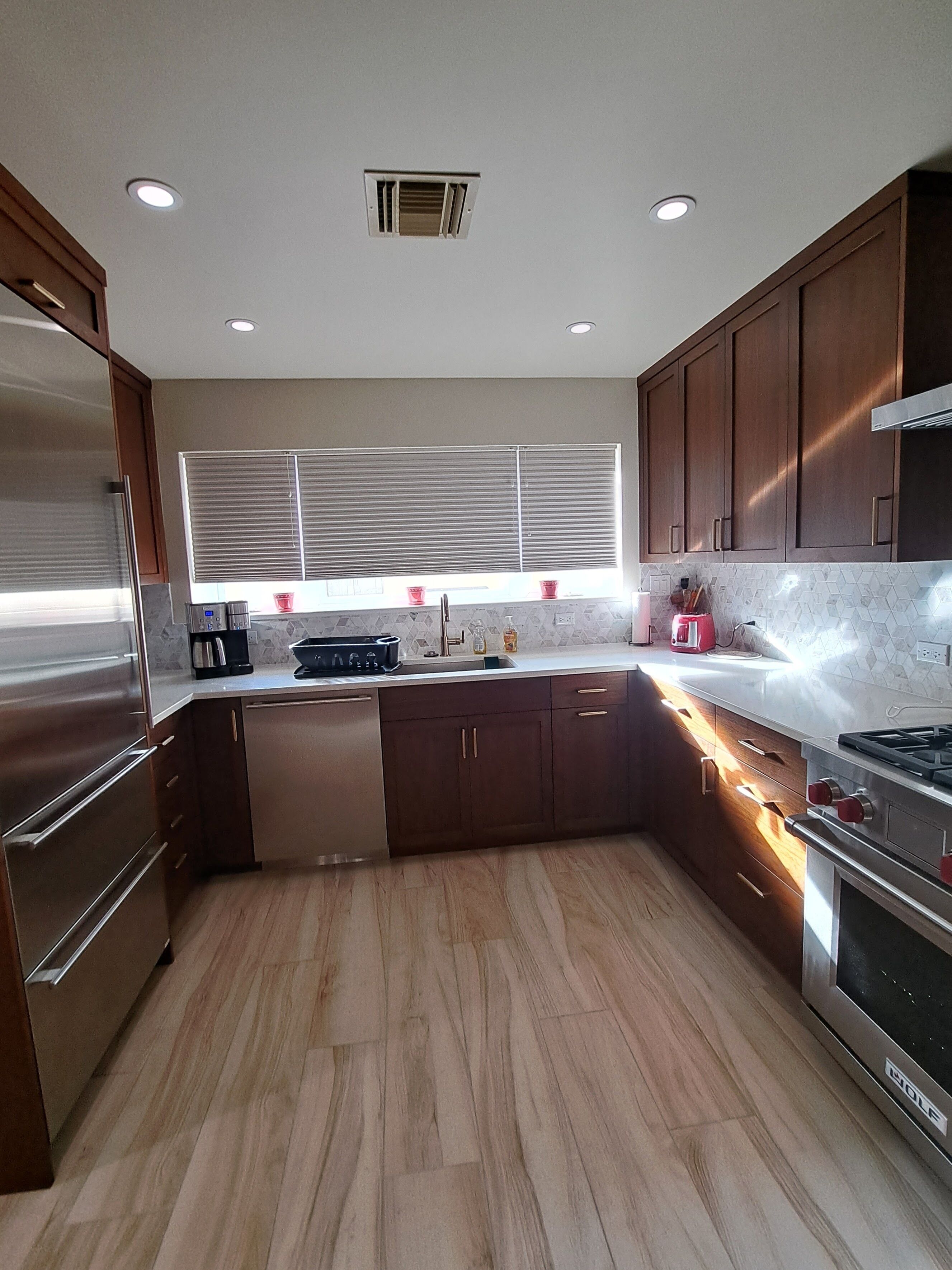 A modern kitchen with wooden cabinets, a stainless steel refrigerator, a built-in oven, and a window with blinds.