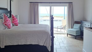 Een slaapkamer met een bed, nachtkastje, stoel en uitzicht op het strand door een groot raam.