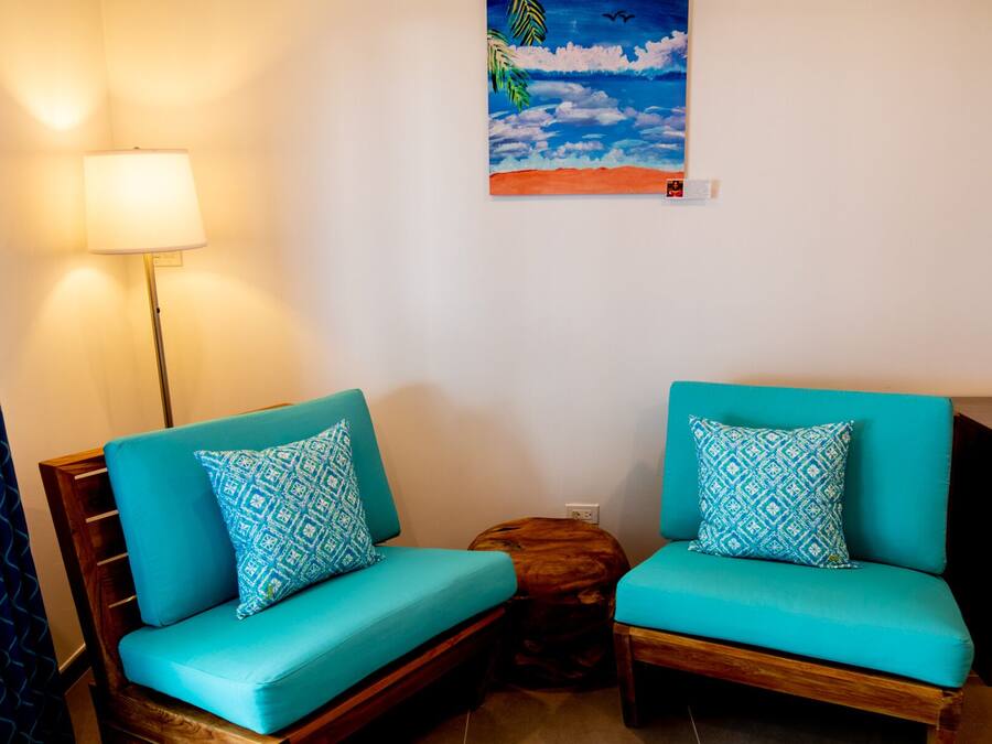 A room with two turquoise sofas with patterned cushions, a wooden side table, a wall-mounted lamp, and a painting of a beach scene.