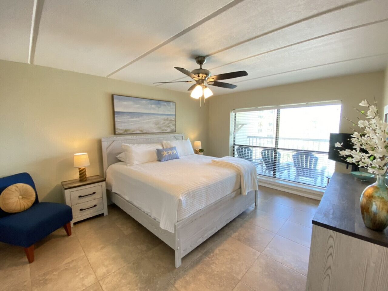 A bedroom with a bed, nightstands, a chair, a ceiling fan, a window, and a balcony with a railing.