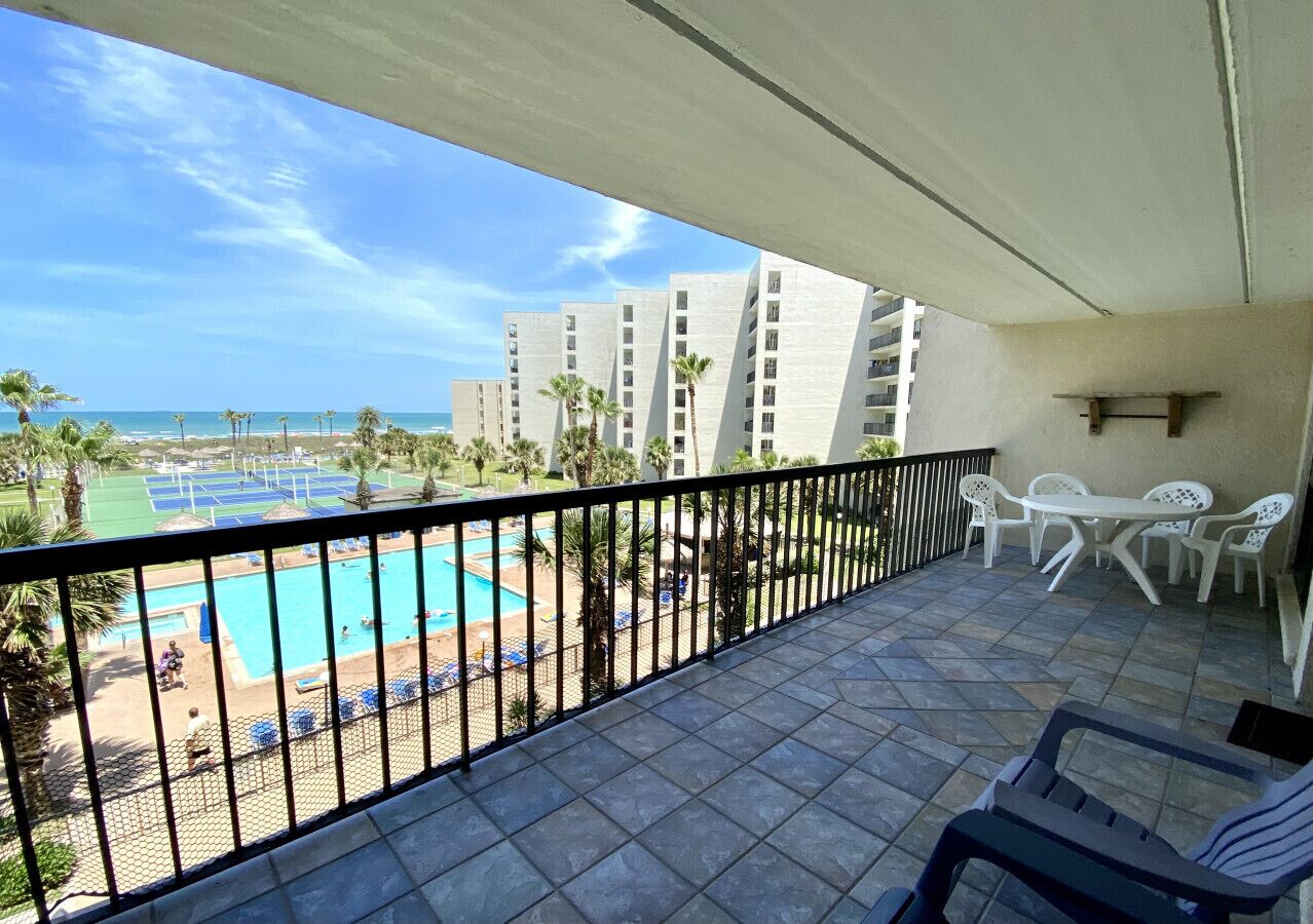 A balcony with a view of a swimming pool and beach, white chairs, and a table.
