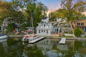 A lakeside property with a dock, a boat, and a house with a deck and outdoor seating area. - Three steps away from the water on beautiful Okauchee Lake! (Okauchee Lake)
