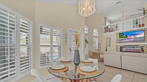 A modern living room with a glass dining table, white chairs, a large TV, and a balcony with white shutters. - Welcome to Alena's Place! Only minutes away from Disney! (Kissimmee)
