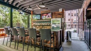 Un espace bar extérieur avec des chaises en osier et un comptoir en bois, agrémenté d’une enseigne ancienne et d’une vitrine.
