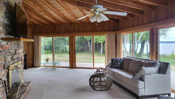A spacious living room with a stone fireplace, a ceiling fan, and large windows offering a view of a lake and forest. - Lake Michigan Retreat (Menominee)