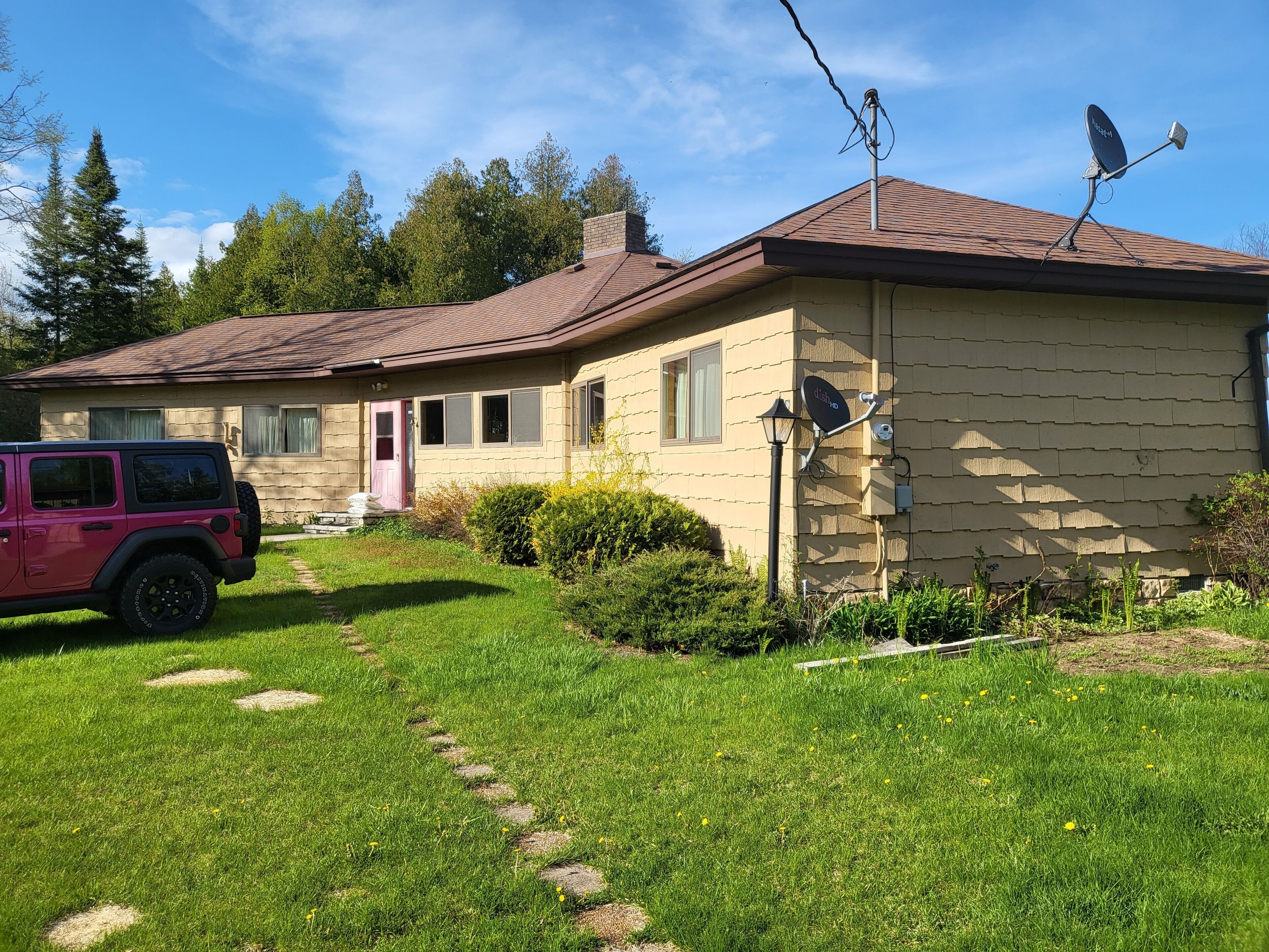A single-story house with a paved driveway leading to the front door, a satellite dish on the roof, and a pink SUV parked in the driveway.